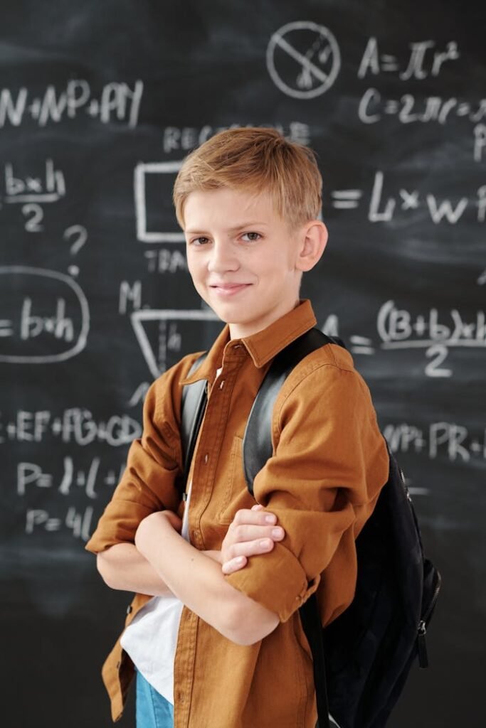 pexels photo 5212324 Smiling elementary student standing in classroom with math equations on blackboard.